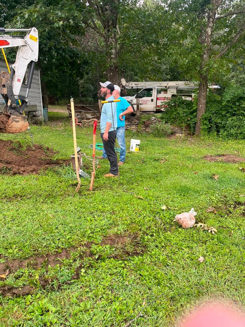 Two crew members standing over a freshly dug well head in a yard, with the lettered service truck and excavator behind them
